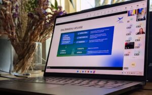 Uz galda pie loga novietots portatīvais dators ar atvērtu tiešsaistes sanāksmi; ekrānā redzama prezentācijas slaids par dalībnieku atlasi un labajā pusē – videokonferences dalībnieku logi. Blakus datoram vāzē redzami kaltēti ziedi.