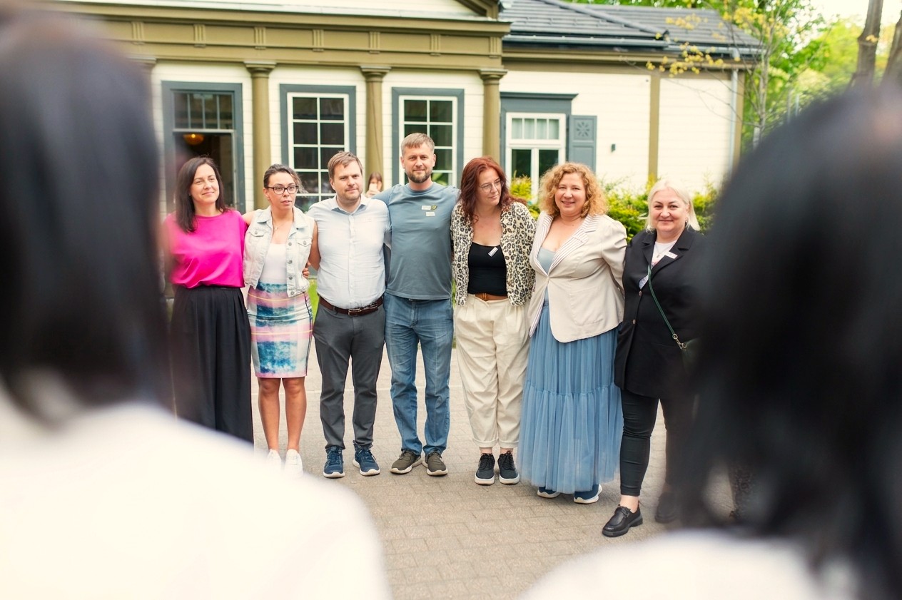 Cilvēki stāv kopīgi plecu pie pleca vienā līnijā, aplikuši rokas viens otram ap pleciem.